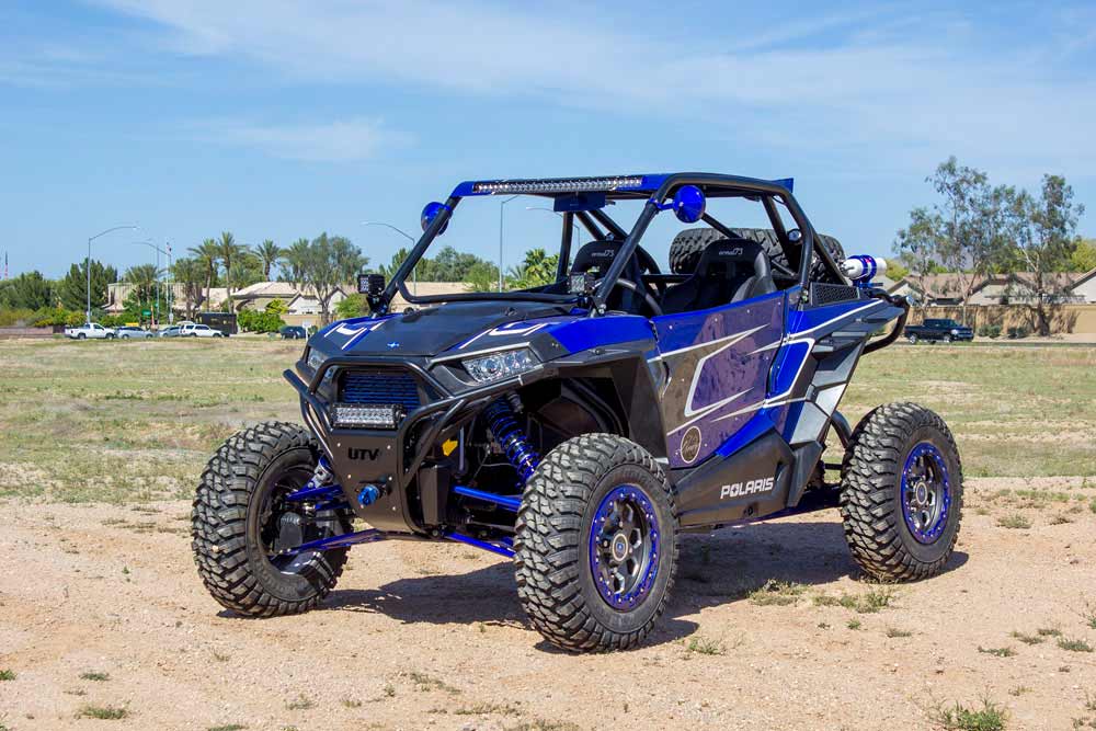 Blue and black Polaris RZR with an aftermarket roll cage, LED light bar, blue anodized suspension links and blue beadlock wheels, parked on a dirt lot